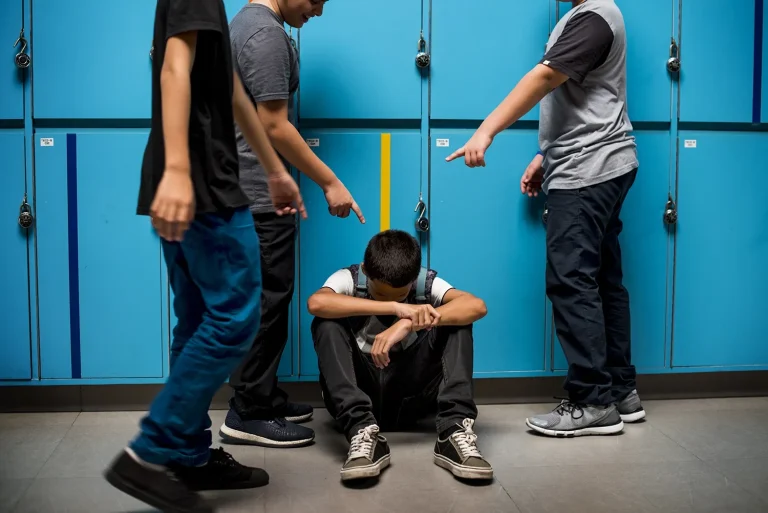 Een jongen zit met zijn hoofd omlaag tegen een rij blauwe schoolkluisjes, terwijl drie medeleerlingen hem omringen en met vingers naar hem wijzen – een confronterend beeld van pesten op school.